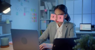 Masada oturan Asyalı genç iş kadını geceleri ofiste dizüstü bilgisayar başında uyuyakalıyor. Yorgun bir kadın. Gözlerinin üzerinde iki post-it var. İş yerinde uyuyan yüz üzerine yapışkan notlar..