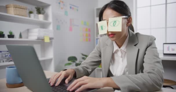 デスクに座っている若いアジアのビジネスマンは オフィスのノートパソコンの上で疲れた睡眠を過労しました 彼女の目の上に2つのポストItと疲れ燃え尽きた女性 職場で眠っている顔に接着剤ノート — ストック動画