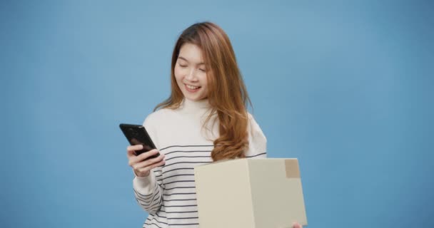 若い陽気なアジアの女性はカジュアルセーターホールドパッケージ小包ボックスを着用し スマートフォンは青の背景に隔離されたオンラインショッピングスタンドをお楽しみください 宅配便と配送サービスのコンセプト — ストック動画