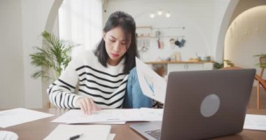 Genç Asyalı bayan serbest olarak dizüstü bilgisayardaki sunum işine ve mutfaktaki şirketle uzaktan görüşmenin finans projesine odaklanıyor. Kız öğrenci internette öğreniyor..