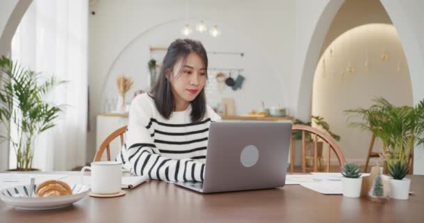 若いアジア女性のフリーランスは 自宅のキッチンで会社とのリモートミーティングのためのアカウントと金融プロジェクトのためのラップトップでのプレゼンテーション作業に焦点を当てています 女の子学生は自宅でオンラインで学ぶ — ストック動画