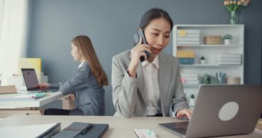 Genç Asyalı iş kadınları cep telefonlarıyla konuşuyor ve iş yerindeki ofis masasında oturarak iletişim kuruyor. Uzak webinar, internet teknolojisi, yaşam tarzı sosyal uzaklık ve Coronavirüs 'ten sonra çalışma.