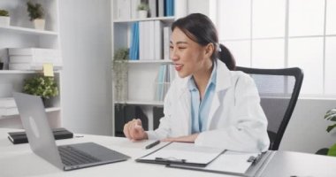 Steteskoplu, beyaz üniformalı genç Asyalı doktor. Hastanedeki ya da klinikteki bir hastayla bilgisayarla konuşan video konferans görüşmesi yapıyor. Danışmanlık ve terapi konsepti.