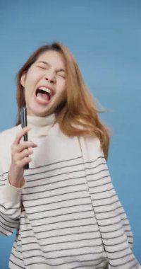 Genç Asyalı bayan karaoke yapan süveter giyip telefonları açık tutup mavi arka planda izole bir şekilde dans ediyor. Gençler evlerinde karantina altında yaşarlar. Dikey ekran.