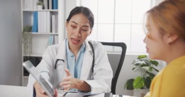 Tablet kullanan beyaz üniformalı genç Asyalı kadın doktor harika haberler veriyor. Sonuçları ya da belirtileri sağlık kliniğinde ya da hastane ofisinde masada oturan kız hastayla tartışıyor..
