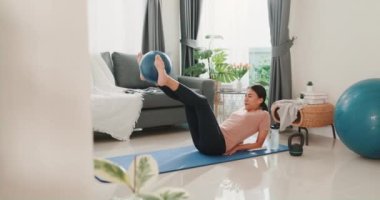 Spor kıyafetleri içinde genç ve çekici Asya 'lı güçlü bir kadın. Oturma odasında spor topuyla ısınıyor. Ev karantina idmanı ve spor..