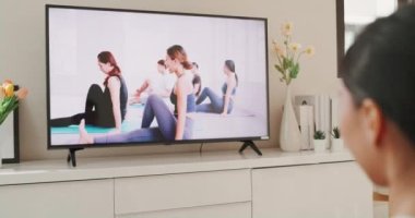 Young attractive Asian woman in sportswear site on mat in the floor virtual fitness class with group of people on a video conference on television practice yoga for beginner in living room at home.
