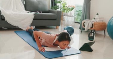 Young attractive Asia woman in sportswear watching fitness online video on tablet practice yoga for beginner in living room at house. Home quarantine workout and fitness exercise without the gym.
