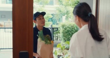 Young Asian delivery man in blue navy uniform knock door home delivery grocery online shopping to woman customer in front of the door and woman accept paper bag at house. Delivery service concept.