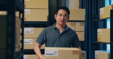 Successful Asian businessman stand front of shelf hold cardboard paper box smiling and looking at camera proud to present of his business in warehouse at night. Startup small business concept.