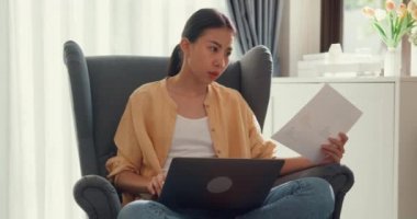Young attractive asia female girl or university student sit on sofa chair with computer laptop check data chart in paperwork on report work in living room at home. Home lifestyle concept.