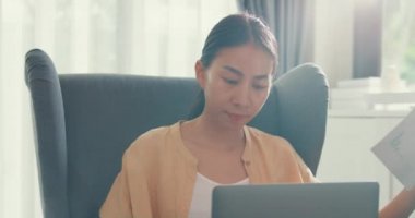 Close-up Young asia female girl or university student sit on sofa chair with computer laptop typing and checking data chart in paperwork on report work in living room at home. Home lifestyle concept.