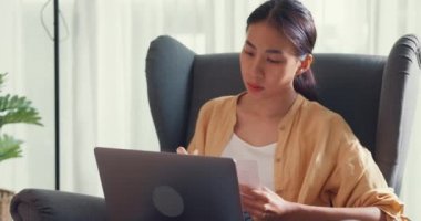 Close-up Young asia female girl or university student sit on sofa chair with computer laptop typing and checking data chart in paperwork on report work in living room at home. Home lifestyle concept.