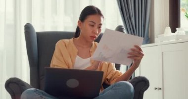 Young attractive asia female girl or university student sit on sofa chair with computer laptop check data chart in paperwork on report work in living room at home. Home lifestyle concept.