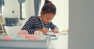 Asian toddler girl with sweater sit in front of desk with notepad use pencil focus on write notebook do homework from online learning course on the weekend at home. Distance online learning concept.
