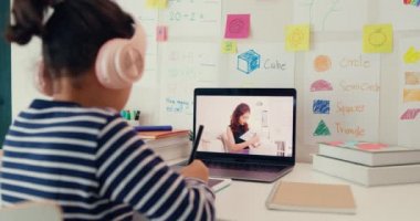 Asian toddler girl with sweater wear headphone sit front of desk with notepad use magic pen focus on writing do homework listen teacher online learning english course from laptop screen at home.