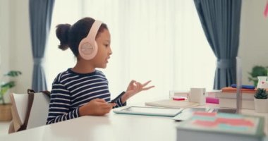 Asian toddler girl with sweater wear headphone sit front of desk with notepad use magic pen focus on writing do homework, finger counting number math online learning course from laptop at home.