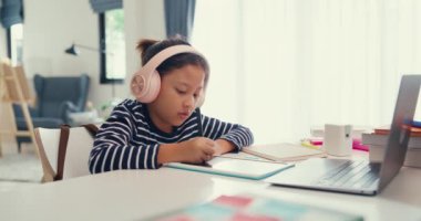 Asian toddler girl with sweater wear headphone sit front of desk with notepad use magic pen focus on writing do homework, finger counting number math online learning course from laptop at home.
