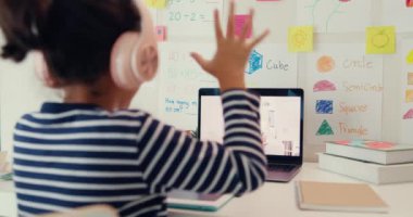 Asian toddler girl with sweater wear headphone sit front of desk with notepad use magic pen focus on writing do homework listen teacher online learning english course from laptop screen at home.