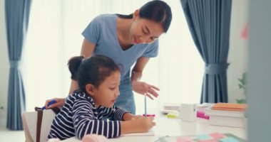 Asian toddler girl with sweater sit in front of desk with notepad use pencil focus on write notebook do math homework with her mother or tutor on weekend at home. Distance online learning concept.