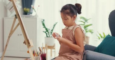 Asian toddler girl with apron sit on sofa chair in front of painting canvas with color palette use paint brush focus on painting abstract on weekend at home. Creative lifestyle for kid concept.