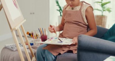 Close-up Asian toddler girl with apron sit on sofa chair in front of painting canvas with color palette use paint brush focus on painting abstract on weekend at home. Creative lifestyle kid concept.