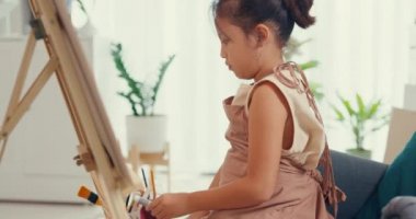 Asian toddler girl with apron sit on sofa chair in front of painting canvas with color palette use paint brush focus on painting abstract on weekend at home. Creative lifestyle for kid concept.