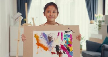 Asian toddler girl with apron stand front sofa chair holding up her art work natural sweet smilling looking at camera show abstract art work in living room at home. Creative lifestyle kid concept.
