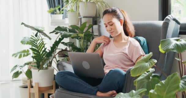 若いアジアの女性は リビングルームの温室で植物でいっぱいの仕事をするためのアイデアを考えるラップトップコンピュータを使用して 翼の椅子に座ってカジュアルな穏やかな座って着用してください コロナウイルスと家にいなさい家のコンセプトから仕事 — ストック動画