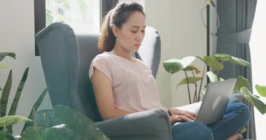 Genç Asyalı bir kadın kanat koltuğunda rahat bir şekilde otururken dizüstü bilgisayar kullanıyor ekran daktilo raporuna odaklanıyor ve oturma odasında bitki yetiştirmekten yoruluyor. Ev kavramından çalış.