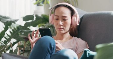 Yakın plan, sıradan kulaklık takan genç Asyalı kadın kanat sandalyesine oturur, gözlerini kapatır telefon kullanır evdeki oturma odasında sakin bir müzik dinler. Konforlu ev iç mekanlar, şehir ormanı konsepti..