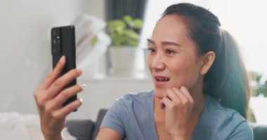 Yakın plan genç Asyalı kadın kanepeye günlük kıyafet giyer. Akıllı telefon kullanır. Video çekip merhaba der, aile ve arkadaşınla mutlu bir şekilde konuşur ve evdeki ev bitkilerinde gülümserdi..