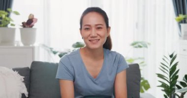 Genç Asyalı kadın rahat bir şekilde kanepede oturup kameraya bakıyor. Merhaba demek için el kaldırıyor ve aile ve arkadaşlarıyla konuşuyor. Mutlulukla gülüyor..