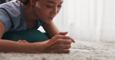 Yakın plan mutlu Asyalı üniversiteli kadın halı döşemesine uzanmış akıllı telefon kullanarak evdeki oturma odasında online ürün alışverişi yapıyor. İç mekan konseptiyle iç mekan..