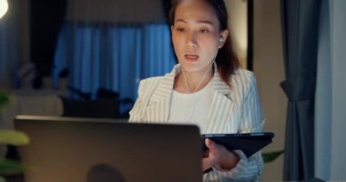 Genç Asyalı iş kadını dizüstü bilgisayarlı resmi oturma masasında otururken kulaklık kullan, internetteki görüşme videosu kullan. Tablet mevcut finans raporunu evdeki oturma odasında sakla. Fazla mesai konsepti.