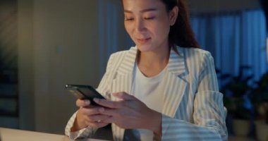 Yakın plan mutlu Asyalı iş kadını resmi kıyafetlerini giyer. Masanın önüne dizüstü bilgisayarla oturup akıllı telefondan mesaj yazar. Evde rahat bir oturma odasında dinlenir. Fazla mesai konsepti.