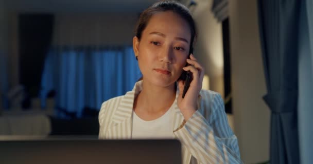 年轻而快乐的亚洲女商人穿着正式的衣服 带着笔记本电脑和平板电脑坐在办公桌前 晚上和同事在舒适的客厅里通电话 加班概念 — 图库视频影像