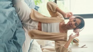 Asyalı bir kız beyaz kremalı pijama giyer. Pembe kulaklık takar. Pofuduk rahat yatağında mutlu bir şekilde dans eder. Dikey Ekran.