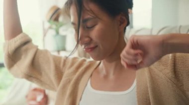 Yakın plan Asyalı kız, rahat yatağında beyaz krem rengi pijama hırka giyer. Uzun bir tatil için uzun bir süre mutlu bir şekilde dans eder. Sabahın köründe, pencereden yatak odasında. Kadın sabah titreşimleri konsepti.