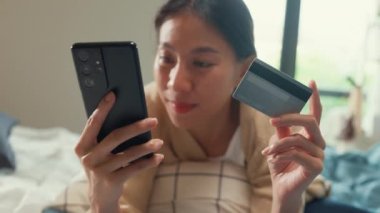 Mutlu, gülümseyen, beyaz krem pijamalı bir Asyalı kız üniversitesi. Kabarık, rahat bir yatağa uzan. Kredi kartı kullan..