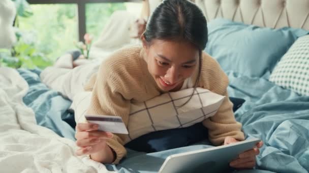 白いクリーム色のパジャマを着た幸せな笑顔のアジアの女子大生は 自宅の寝室でデジタルタブレットでクレジットカードをオンラインショッピングに成功した支払いを使用してふわふわの快適なベッドの上に横たわっていました 室内生活概念 — ストック動画