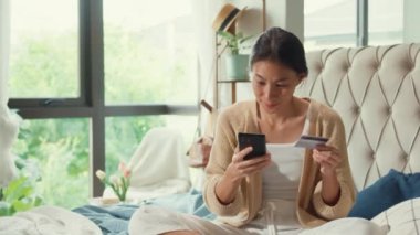 Mutlu gülümseyen Asyalı kız üniversitesi beyaz krem pijamalarıyla kabarık konforlu yatağa uzan kredi kartı kullan online alışveriş kartı kullan yatak odasındaki kameraya bakarken heyecanla cep telefonuna başarılı bir ödeme yap.