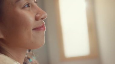 Yakın plan Asya manzaralı, krem pijamalı, beyaz perdeli mutlu kız uyanıyor. Yatakta taze, dışarı bakıyor. Yatak odasının penceresindeki sabah ışığını hissediyor. Kadın yaşam tarzı konsepti.