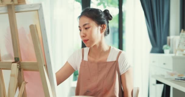快乐的年轻亚洲女性穿着围裙 手握调色板 在家里舒适的车间里用画布在画布上创作艺术品 当代画家摘要现代艺术 创造力与人的概念 — 图库视频影像