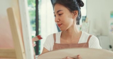 Mutlu genç Asya kadınları palet renginde önlük giyer tuvale fırça boyası sürer evdeki rahat atölyede sanat çalışması yapar. Çağdaş Ressam Soyut Modern Sanat, Yaratıcılık ve İnsan Konsepti.