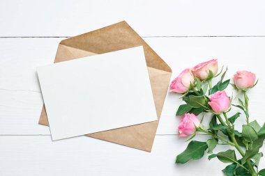 Zarflı boş tebrik kartı modeli ve beyaz ahşap arka planda taze pembe güller, kopyalama alanı olan maketler.