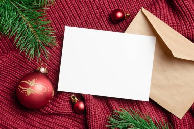 Noel ve Yeni Yıl tebrik kartı maketi zarflı ve kırmızı toplar kırmızı örülmüş arka planda çam ağacı dallarıyla süslenmiş.
