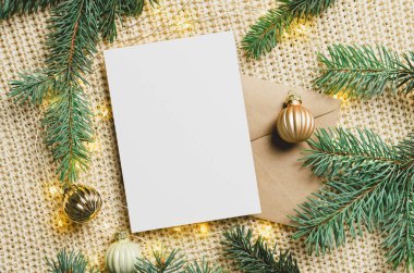 Zarflı Noel tebrik kartı maketi, şenlikli süslemeler ve örülmüş arka planda ışıkları olan köknar ağacı dalları.