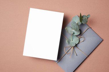 Invitation or greeting card mockup with envelope and natural eucalyptus twig on beige background. Empty card with copy space.