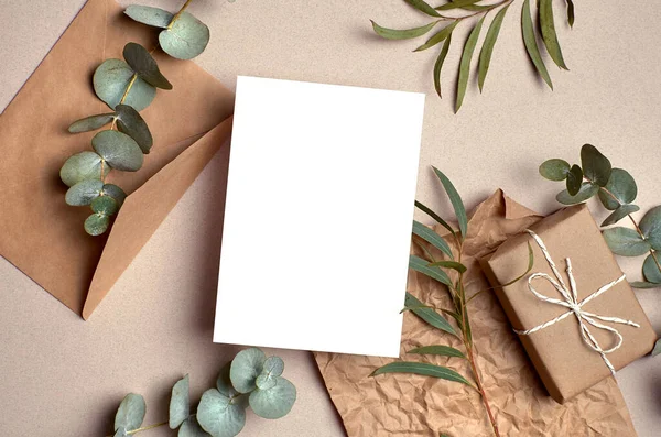 Greeting card mockup with gift box, envelope and natural eucalyptus twigs on paper background. Card mockup with copy space.